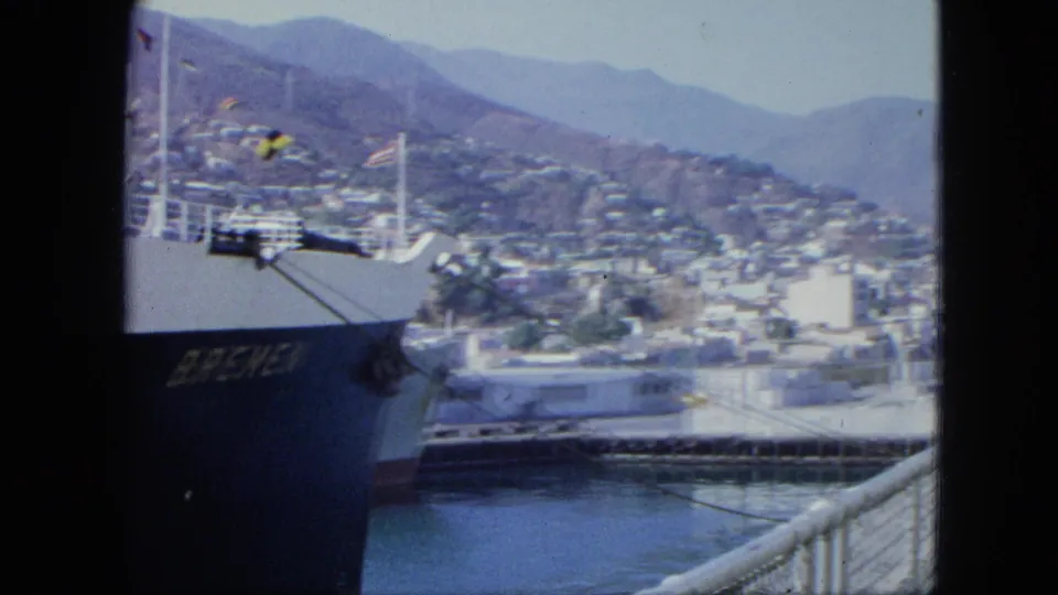 The ship BREMEN is docked at the port, with flags, harbor water, pier railings, and the Venezuelan hillside city in the background.