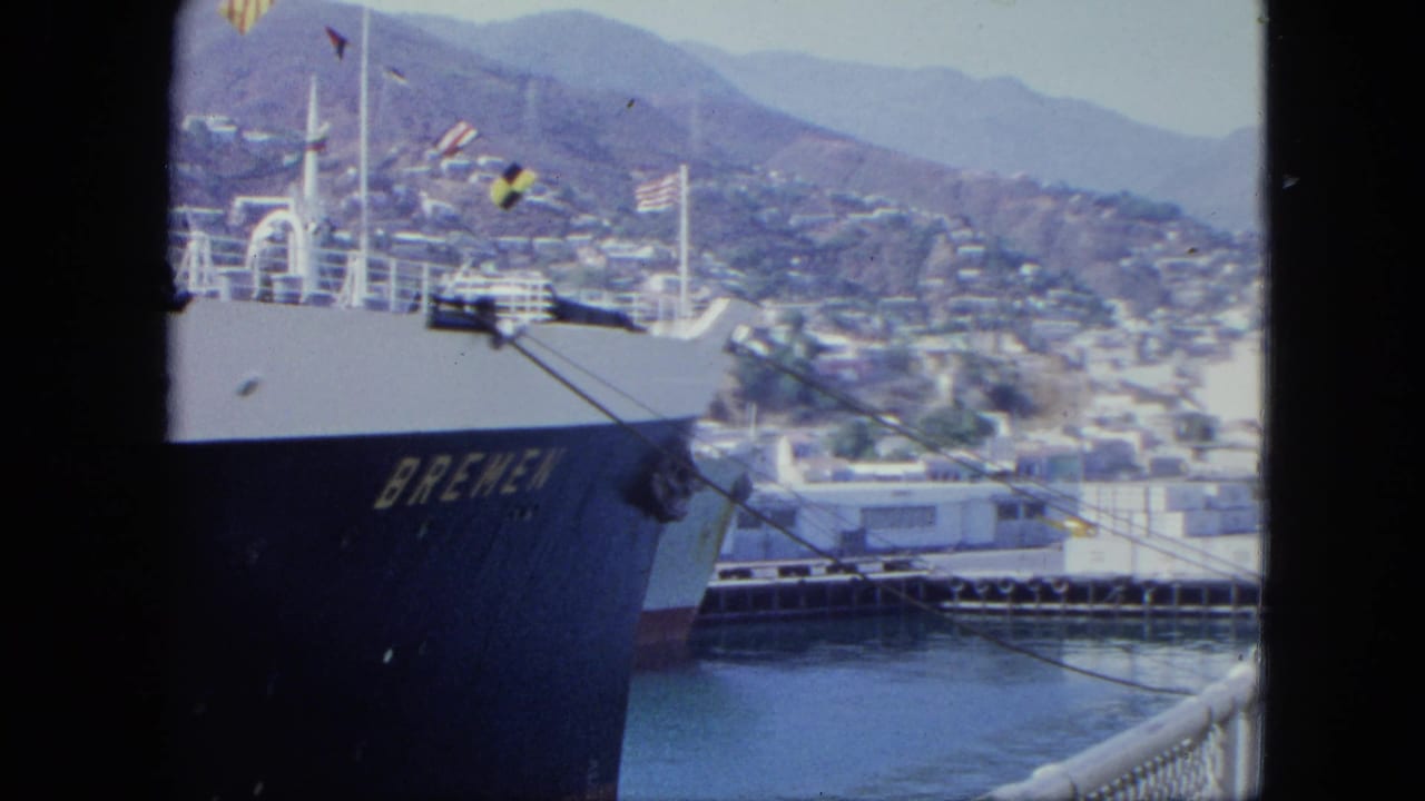 1964 8mm frame from Venezuela showing the Bremen hull, white ship railings, flags, dock structures, water, and a hillside coastal city.