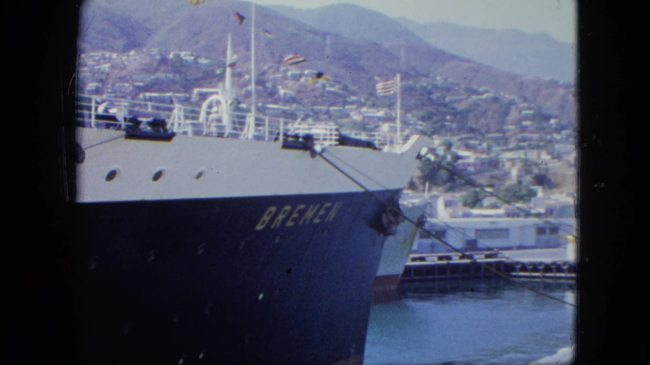 Ship bow and white deck structures fill the foreground with flags and a hillside city in the background.