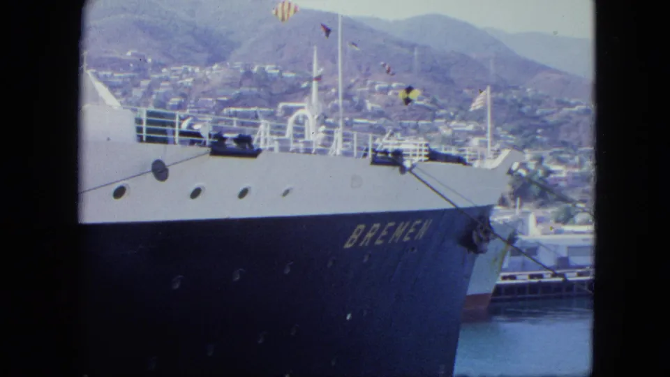 1964 8mm frame in Venezuela showing the side and bow of the Bremen, portholes, railings, flags, harbor water, and hillside city buildings.