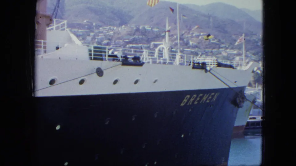 Final 1964 Venezuela frame of the Bremen docked at a coastal port, with visible hull lettering, ship railings, flags, water, and mountain cityscape.