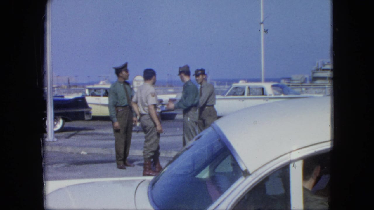 Four uniformed men talk beside parked cars near boats on a bright waterfront sidewalk.