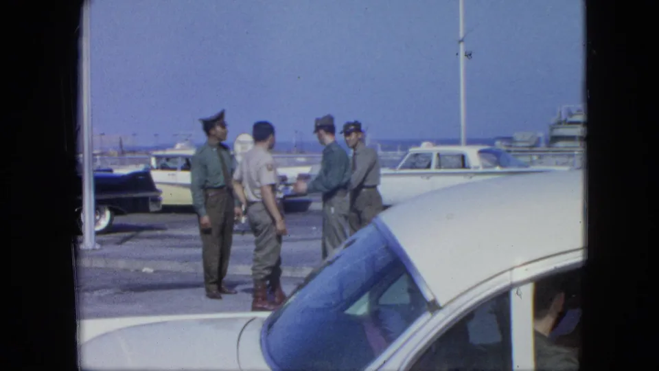 Four men in service-style clothing stand in conversation near parked cars and boats in 1960s 8mm home-movie footage.