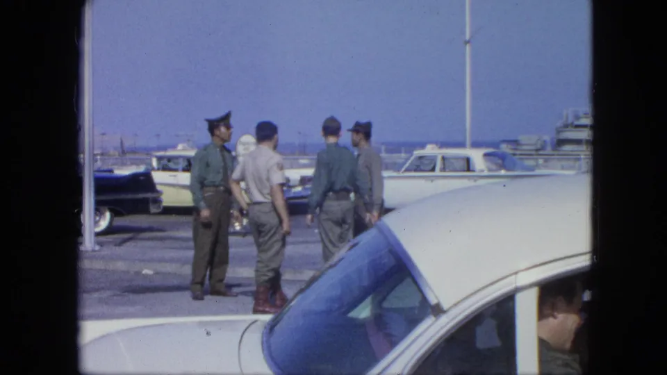 A small group of uniformed men converse beside a white car at a waterfront, with boats and parked vehicles in the background.