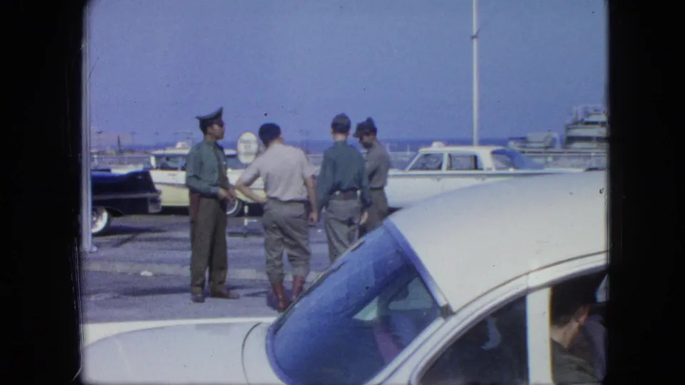 One uniformed man gestures toward the group as the others stand near a parked car and waterfront boats.