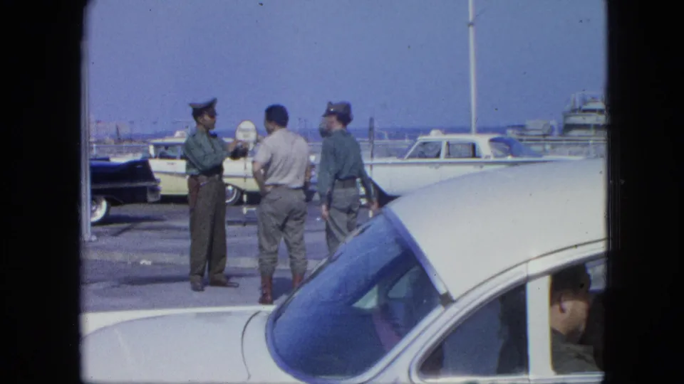 The dockside group continues talking near a white car, with service-style uniforms, parked cars, and boats visible behind them.