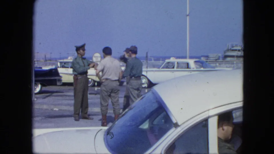 A 1964 8mm frame shows uniformed men and a civilian conversing beside classic cars near a dock or marina area.
