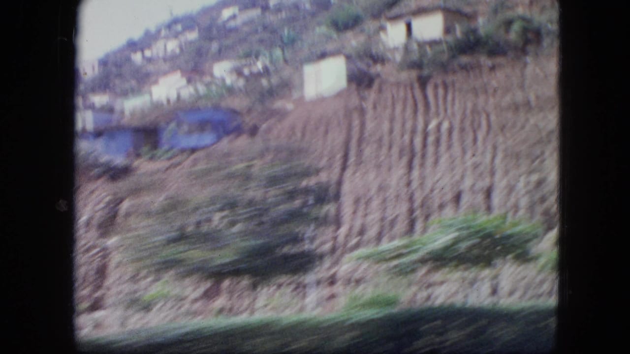 Blurred view of a steep hillside with terraced soil and scattered buildings near water.