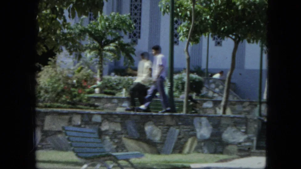 Two people walk through a planted area beside a stone wall, benches, trees, and a light-colored building.