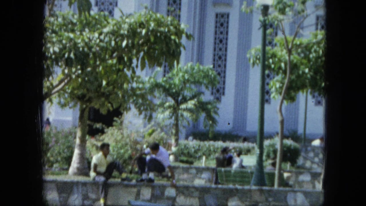 1964 Venezuela 8mm frame showing visitors near a stone garden wall, trees, and part of a white church-like building.
