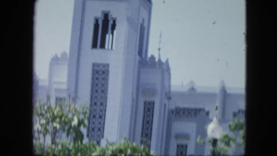 1964 Venezuela home-movie view of a white church-like tower with geometric latticework partly obscured by trees.