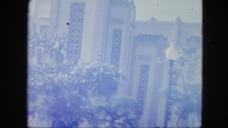 Faded 1964 Venezuela 8mm frame showing the white church-like facade softened by sun glare, trees, and garden plants.