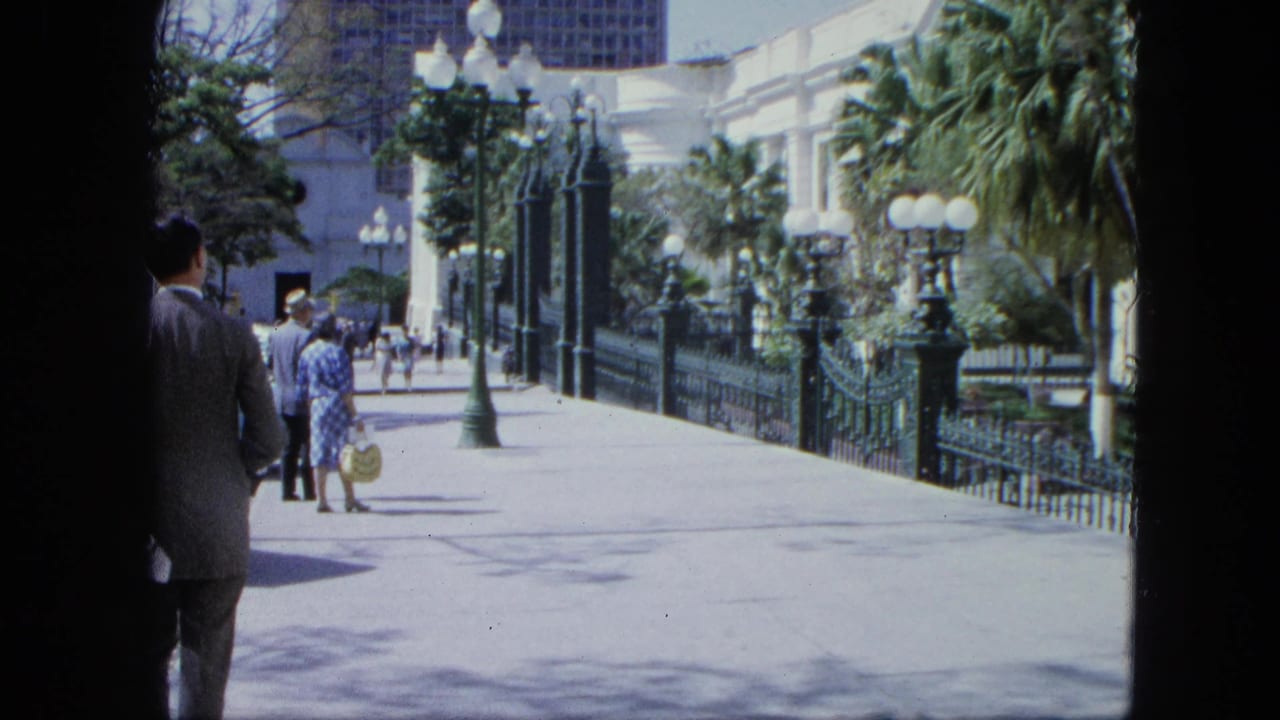 Visitors walk along a sunny promenade beside green iron fencing, palms, and lampposts.