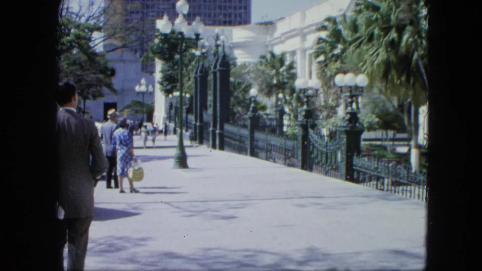 1964 Venezuela home-movie frame of visitors on a bright paved zoo or garden walkway with ornate fencing, palms, and a white building beyond.