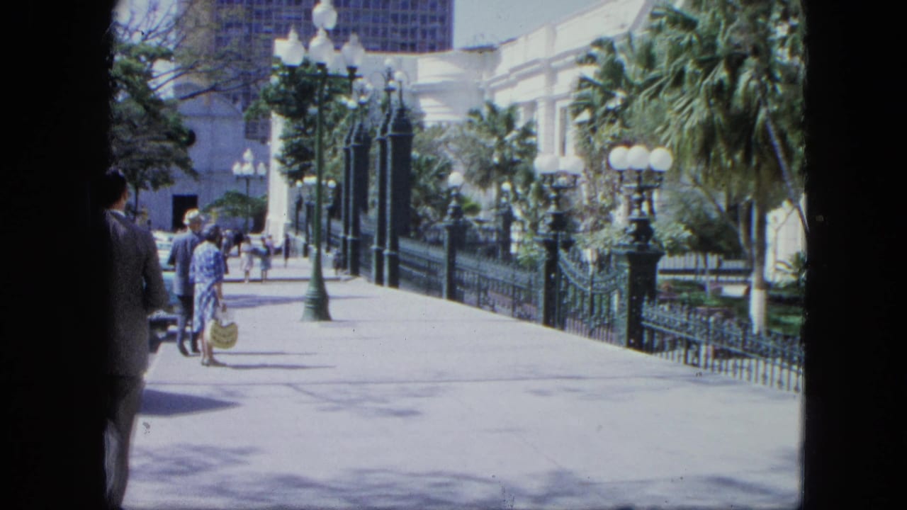 A walkway with visitors, ornate railings, globe lamps, palms, and a white building in bright sun.