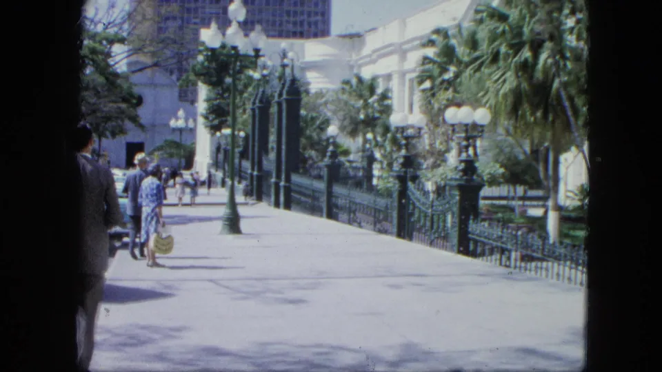 1964 Venezuela home-movie view down a palm-lined zoo or public garden path with visitors near decorative iron fencing and lampposts.