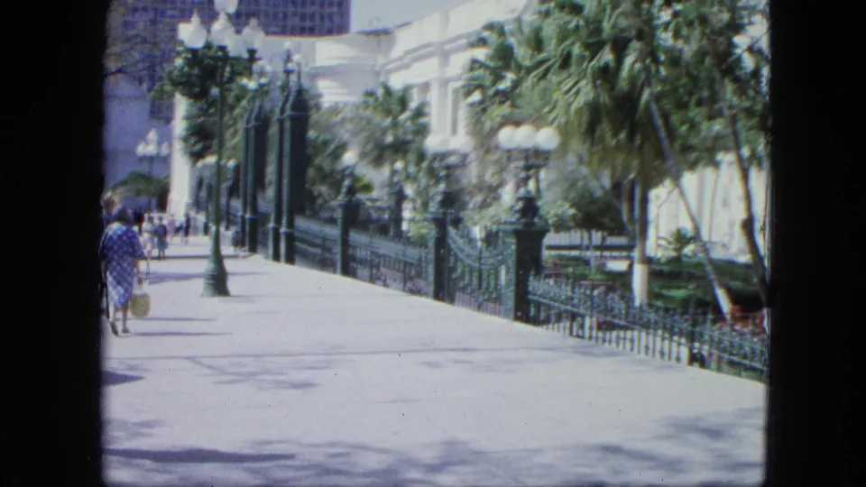 1964 Venezuela frame showing a woman in a blue patterned dress carrying a bag along a bright zoo or garden promenade.