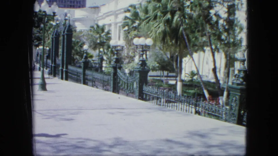 1964 Venezuela home-movie frame of a mostly clear zoo or public garden walkway bordered by ornate fencing, globe lights, palms, and a white building.