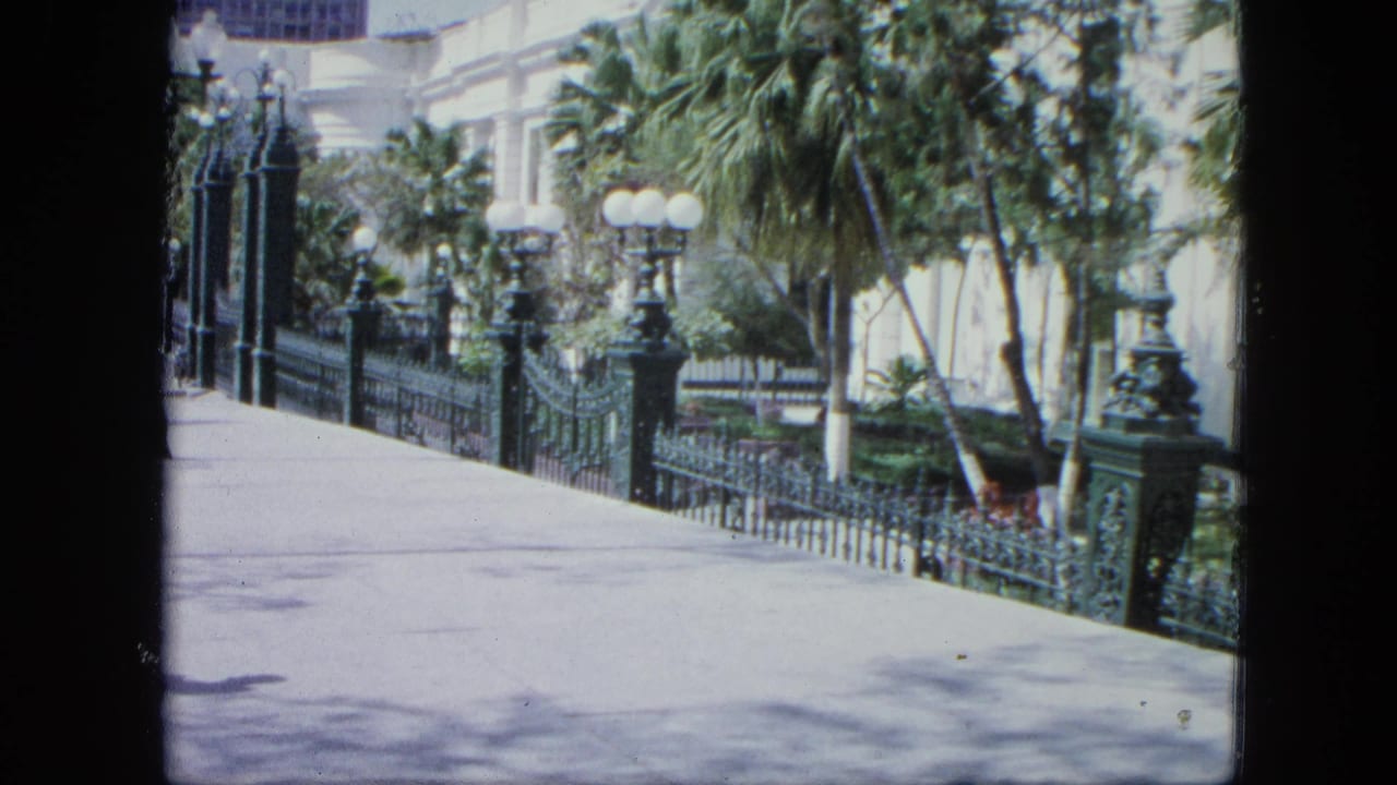 1964 Venezuela 8mm frame showing a bright sidewalk bordered by green iron railings, palm plantings, globe lamps, and a pale exterior wall.