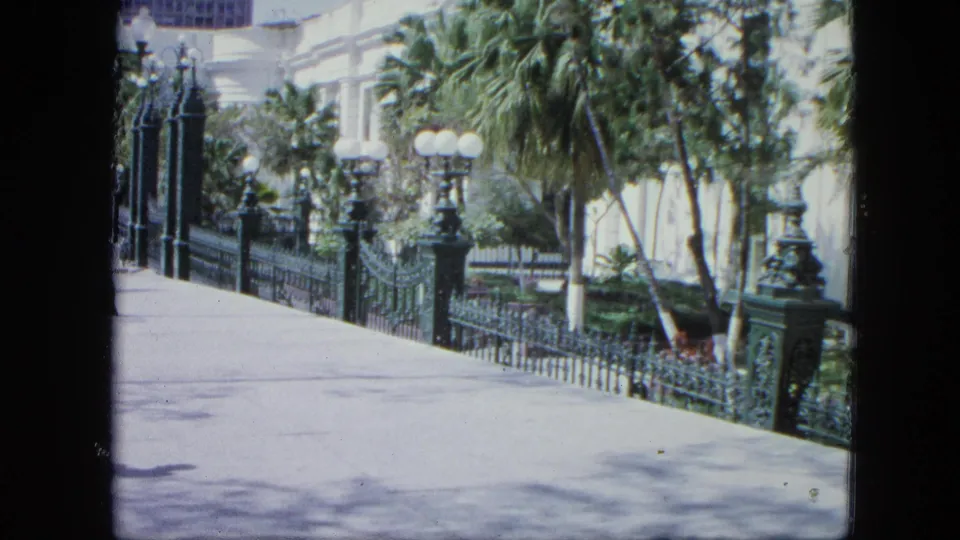 1964 Venezuela 8mm frame showing a bright sidewalk bordered by green iron railings, palm plantings, globe lamps, and a pale exterior wall.