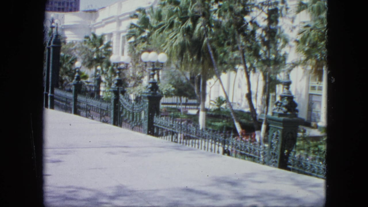 1964 Venezuela travel footage frame with a paved walkway, decorative iron fence, globe lampposts, palms, and a pale building behind foliage.