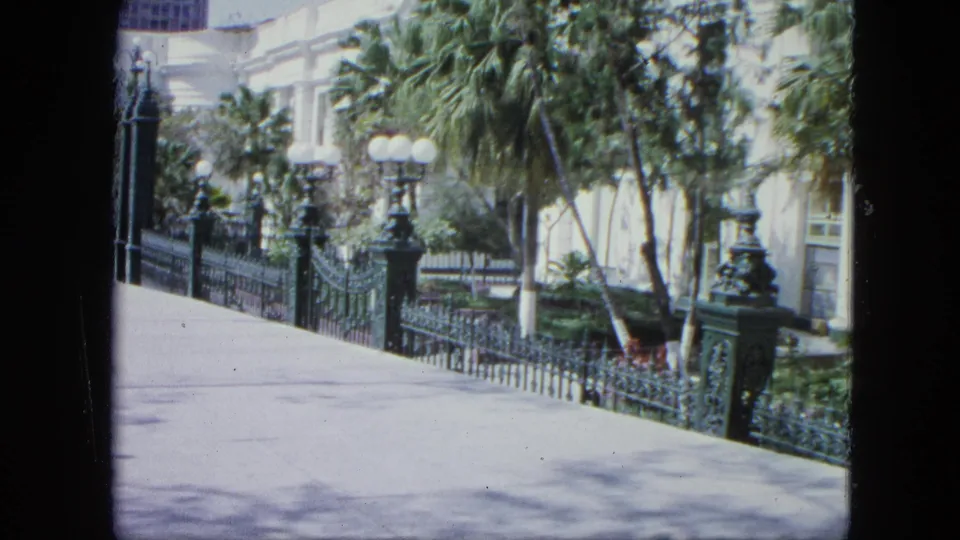 1964 Venezuela travel footage frame with a paved walkway, decorative iron fence, globe lampposts, palms, and a pale building behind foliage.