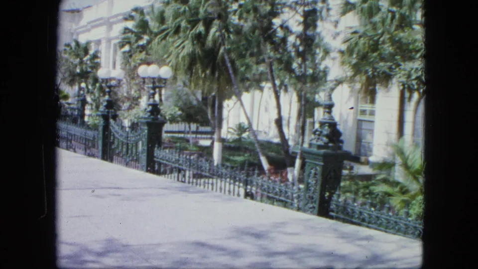 1964 Venezuela 8mm frame from a moving sidewalk view, with ornate green fencing, tropical trees, globe lights, and pale masonry in the background.
