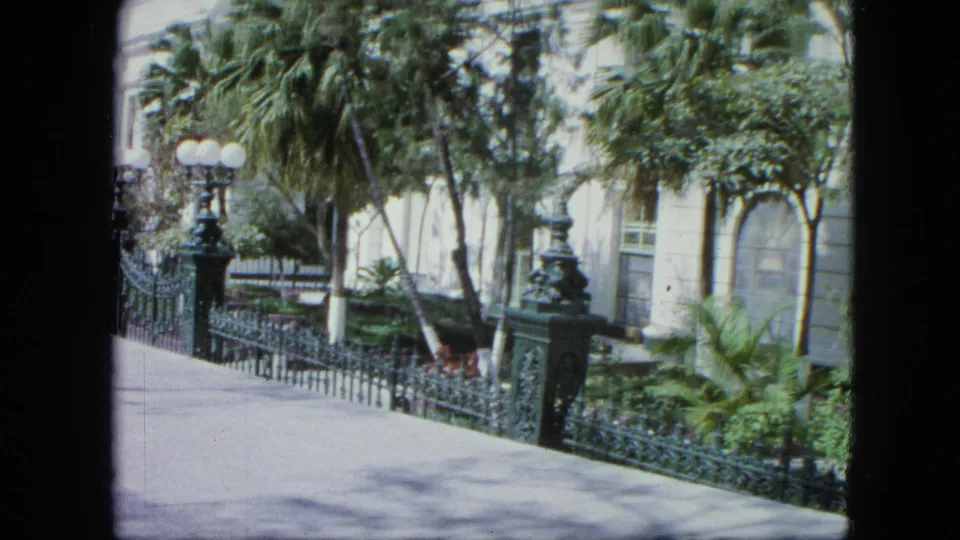 1964 Venezuela frame showing the end of the sidewalk pass, with tropical greenery, iron fencing, globe lamps, and a pale exterior partly hidden by trees.