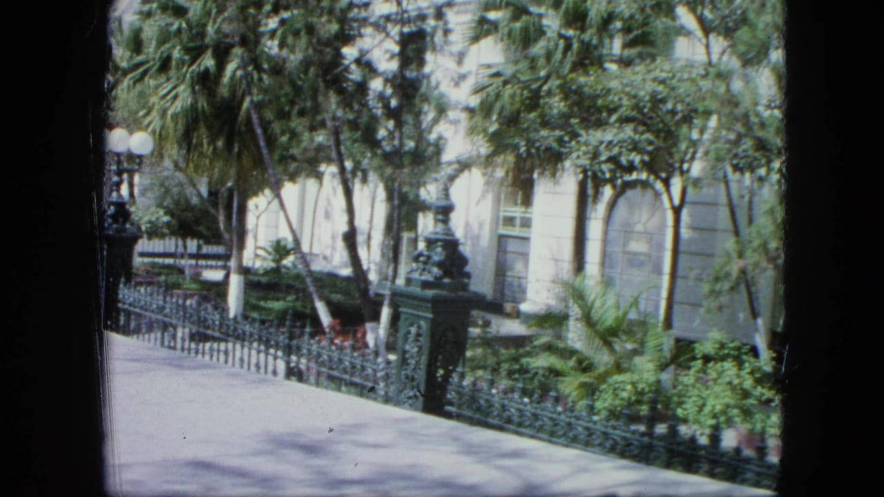 1964 Venezuela home-movie frame showing a white building facade partly obscured by trees, palms, an iron fence, and a globe lamp.
