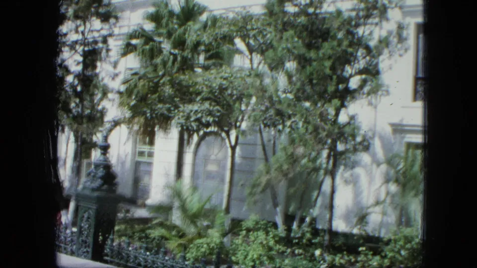 1964 Venezuela home-movie frame showing the camera closer to trees and planting in front of a white building and black iron fence.