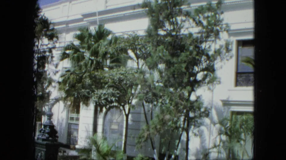 1964 Venezuela frame showing the white building facade, dark window, palms, leafy trees, and dense garden planting in bright sun.