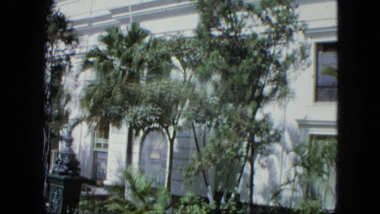 1964 8mm frame of a white colonial-style exterior with arched windows, tropical foliage, iron fencing, and dark frame edges.