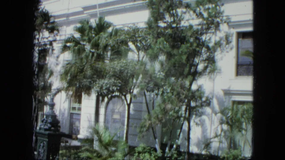 1964 8mm frame of a white colonial-style exterior with arched windows, tropical foliage, iron fencing, and dark frame edges.