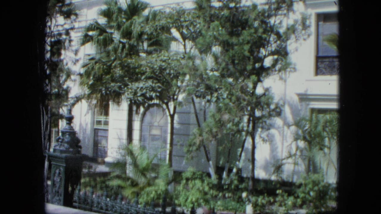 The camera view shifts across a pale exterior in a Venezuela-listed 1964 home movie, with palms, foliage, and a decorative fence visible.