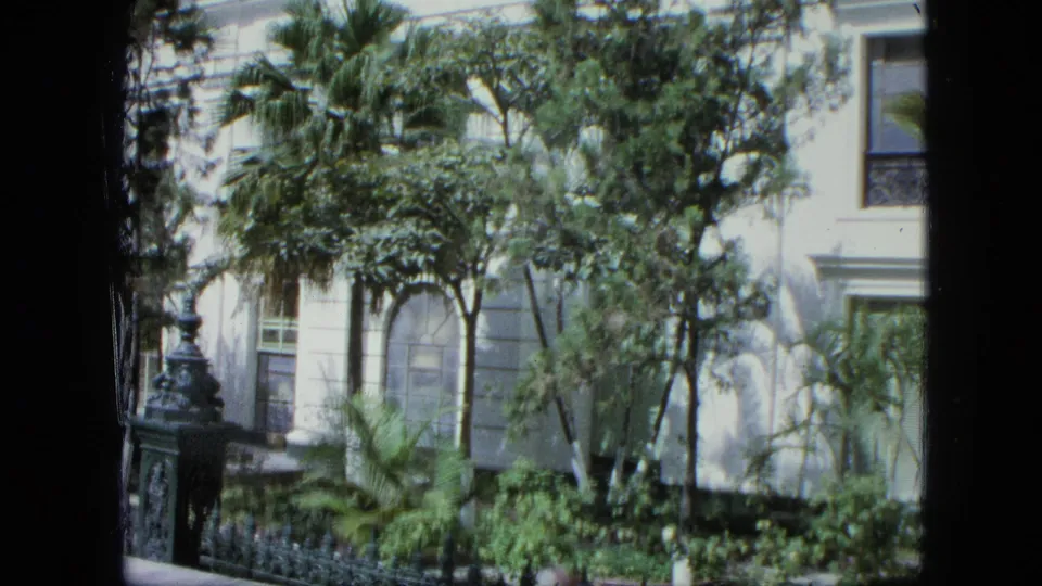 The camera view shifts across a pale exterior in a Venezuela-listed 1964 home movie, with palms, foliage, and a decorative fence visible.