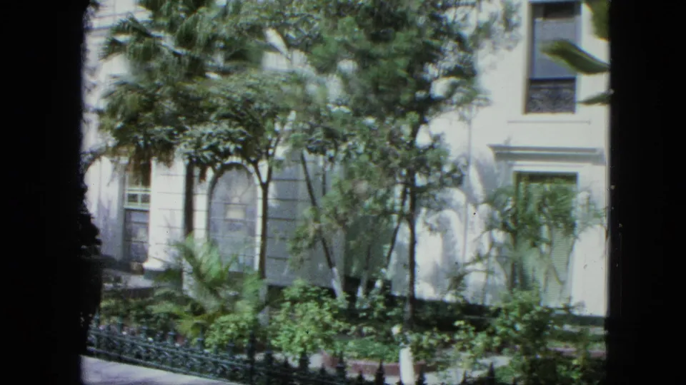 A closer 8mm view shows the white facade, arched window, shrubs, palms, and a dark vertical frame edge during the slow pan.