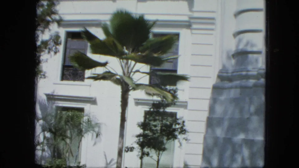 1964 8mm frame capturing the building corner, a central palm, dark shuttered windows, smaller trees, and a curved architectural edge.