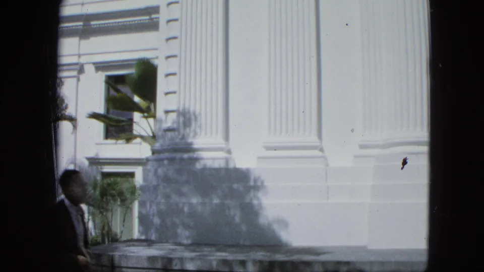 A man in dark clothing walks near a sunlit white building with tall fluted columns, windows, palm fronds, and deep shadows.