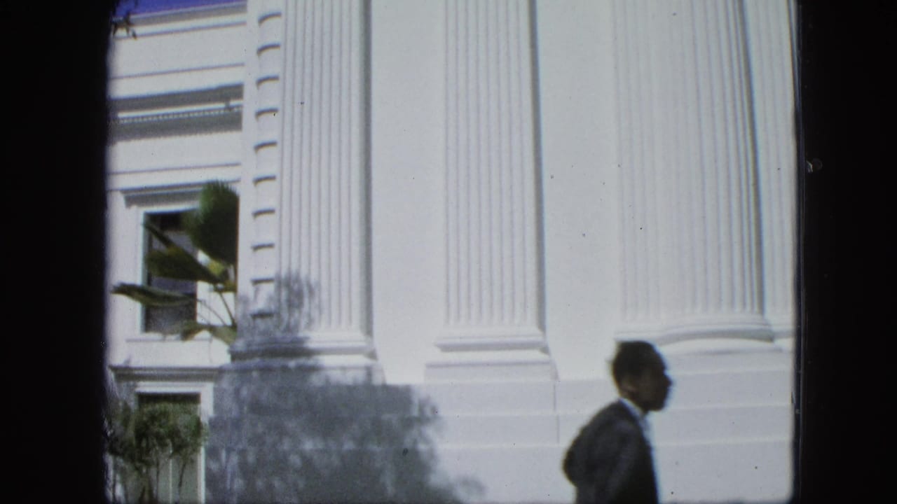 A suited pedestrian crosses the foreground beside a bright facade with fluted columns, a window, palm leaves, and strong sunlight.