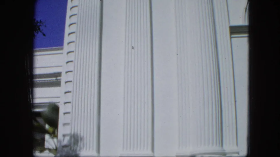 The camera frames the building facade closely, showing tall white fluted columns, a window edge, palm fronds, and blue sky.