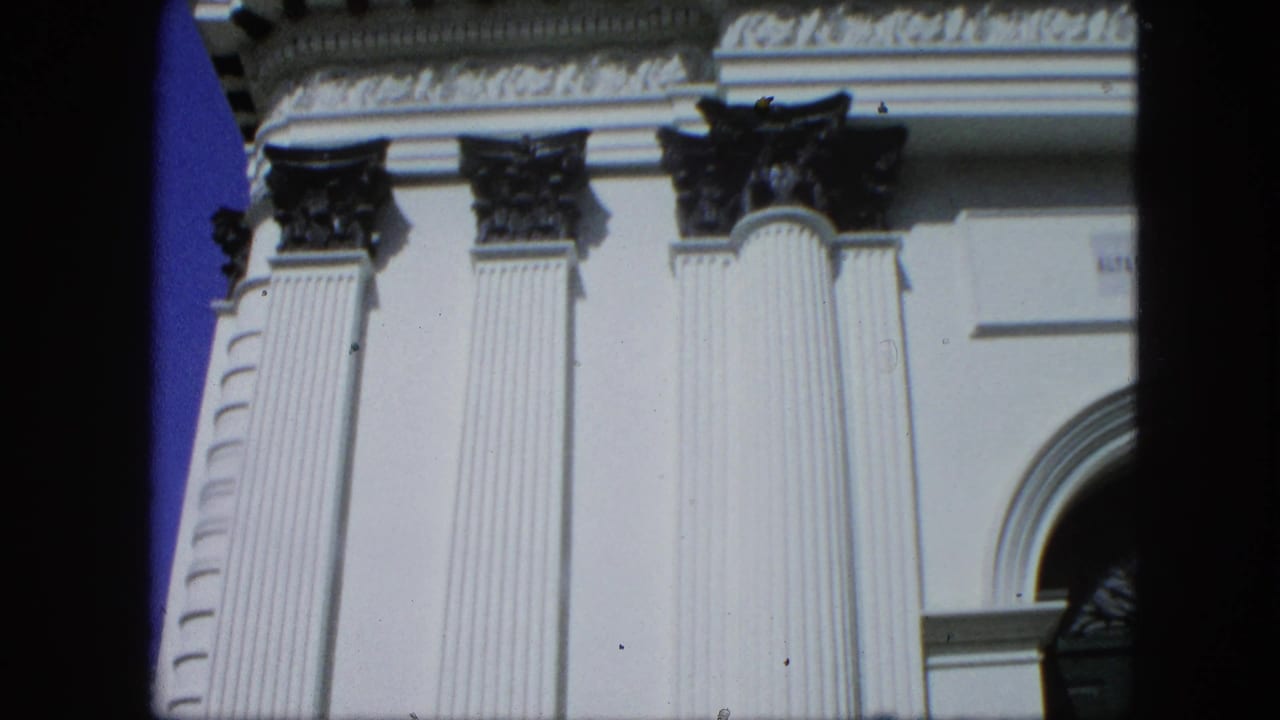 1964 8mm frame showing white fluted columns, dark Corinthian-style capitals, and part of an arched facade, reportedly filmed in Venezuela.