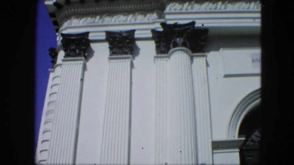 1964 8mm frame showing white fluted columns, dark Corinthian-style capitals, and part of an arched facade, reportedly filmed in Venezuela.