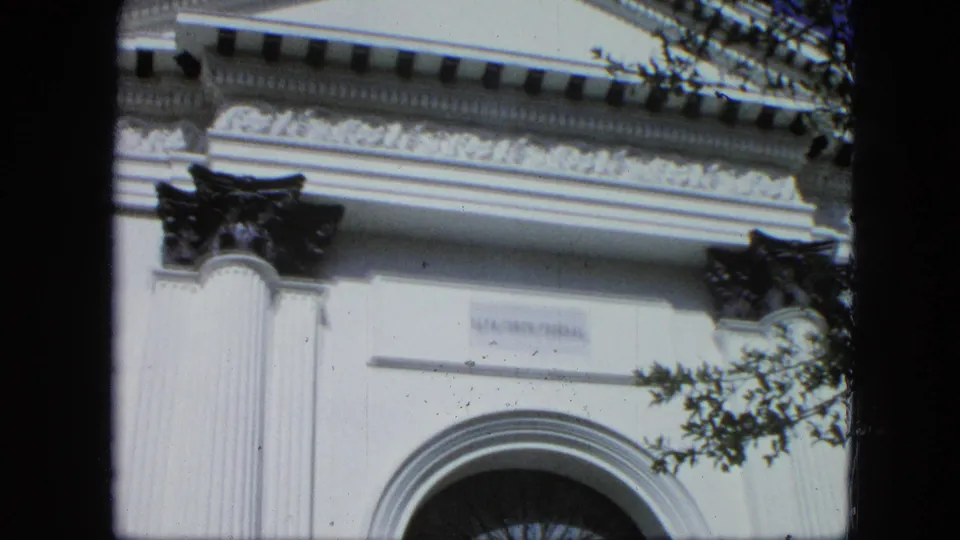 Handheld 8mm view of the building facade with cornice molding, white columns, partial arched doorway, and tree branches crossing the frame.