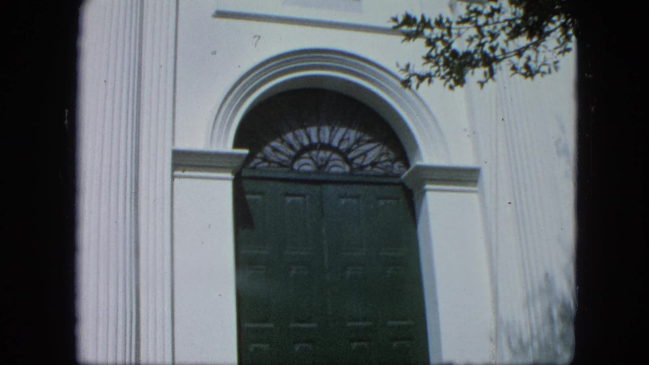 1964 Venezuela home-movie frame of a white neoclassical facade with columns, green double doors, and an arched fanlight.