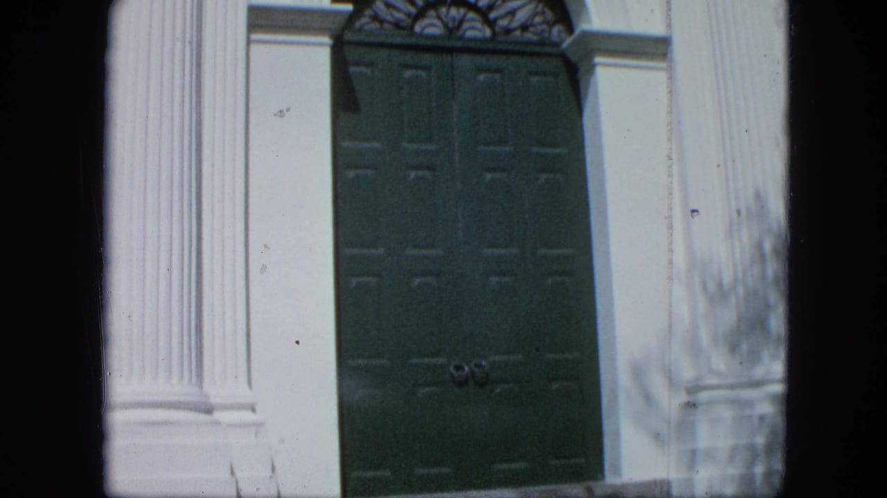 Close 8mm view of tall green paneled double doors set between white columns on a formal building facade in Venezuela.