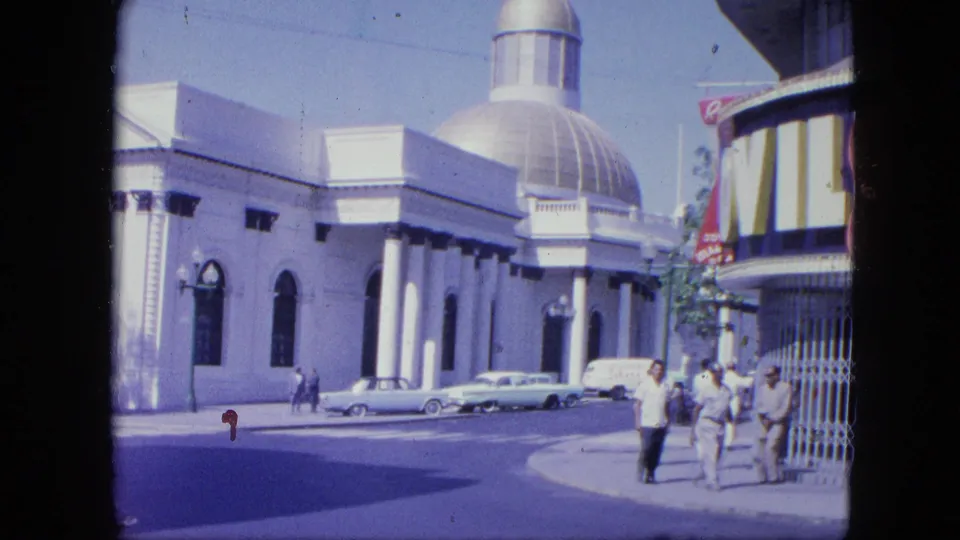 1964 Venezuela street view with a domed public-looking building, parked classic cars, pedestrians, and a curved building edge with partial signage.