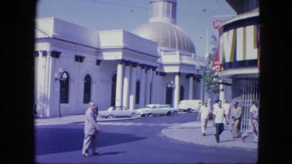 Street-level 1964 Venezuela frame showing adult pedestrians, a domed building, parked classic cars, and partial signage in bright daylight.