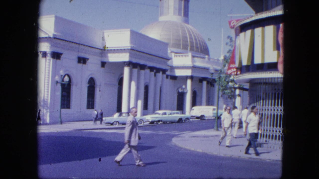 Pedestrians pass a domed columned building beside parked vintage cars on a sunny city street.
