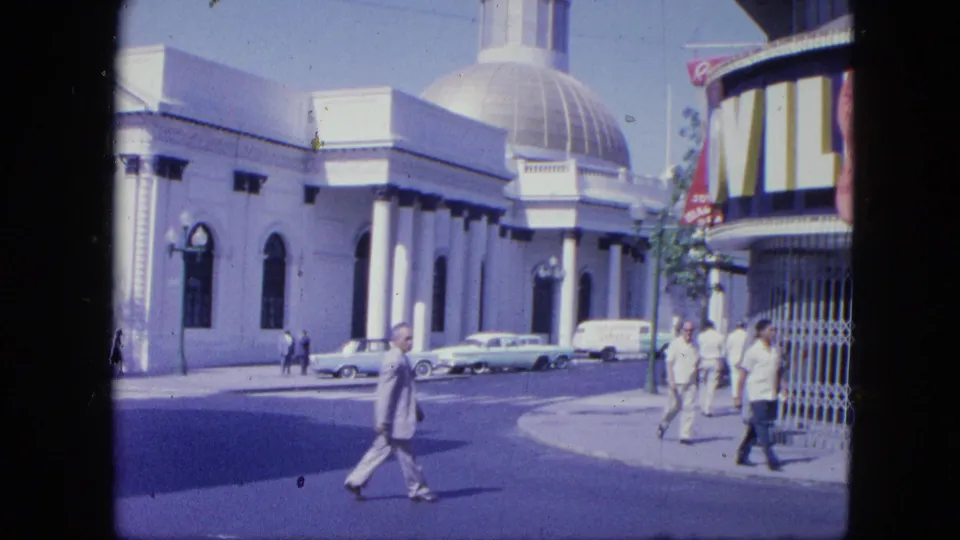 1964 8mm frame of pedestrians near a large domed building with columns, arched windows, parked vintage cars, and partial signage.