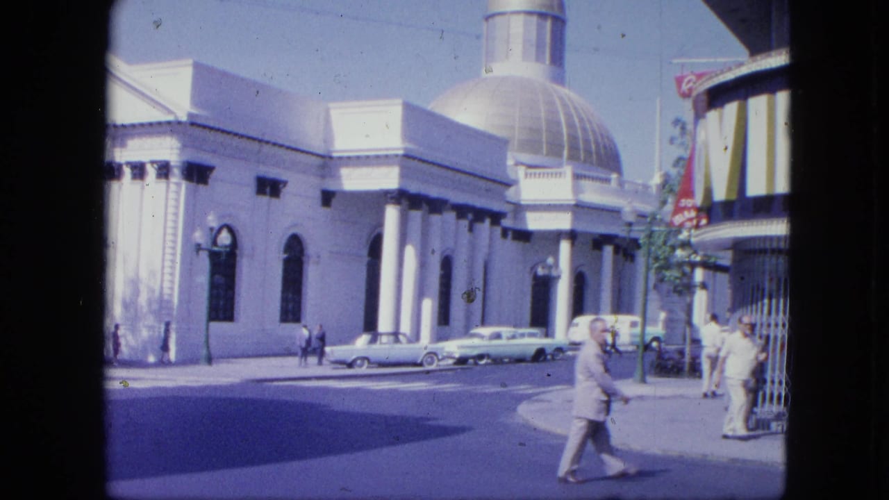 A man crosses in front of a large domed building while other pedestrians stand near signage.